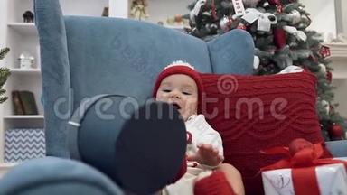 可爱的微笑<strong>宝宝</strong>，带着大<strong>礼</strong>物，坐在一张蓝色的大扶手椅上，在圣诞树附近有一个红色的枕头